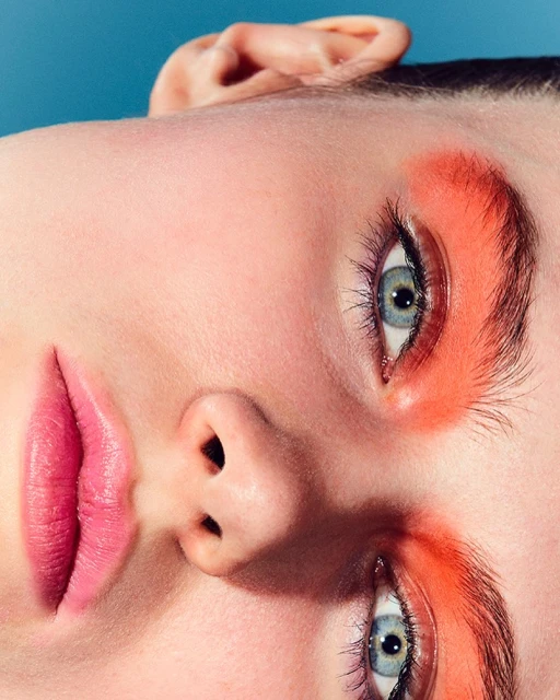 Close-up beauty shot of a model's face featuring vibrant coral eyeshadow and pink lips against a bright blue backdrop for a striking contrast.