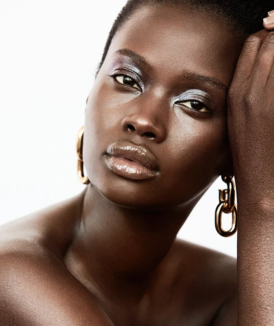 Close-up portrait of a woman with dark skin, wearing gold hoop earrings and silver eyeshadow against a bright, clean white background.