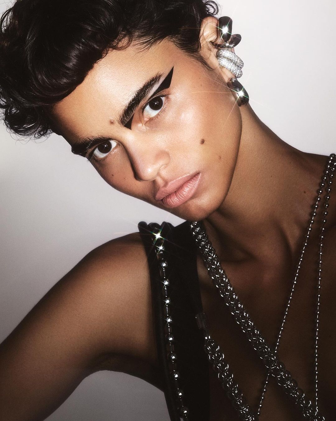 Close-up studio shot of a person with dark hair, bold white graphic eyeliner, silver jewelry, and a sleeveless beaded top.