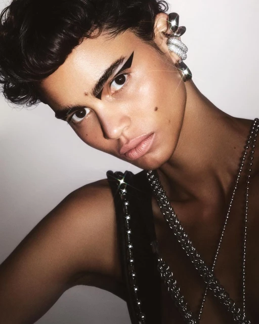 Close-up studio shot of a person with dark hair, bold white graphic eyeliner, silver jewelry, and a sleeveless beaded top.
