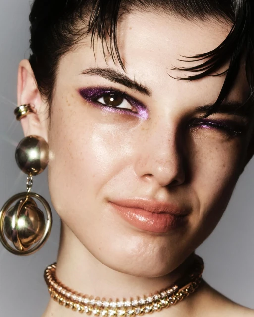 Close-up of a young adult woman with purple eyeshadow and gold jewelry, winking at the camera with a neutral background in a beauty editorial.