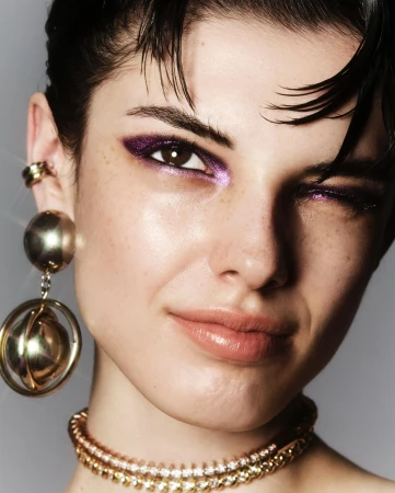Close-up of a young adult woman with purple eyeshadow and gold jewelry, winking at the camera with a neutral background in a beauty editorial.