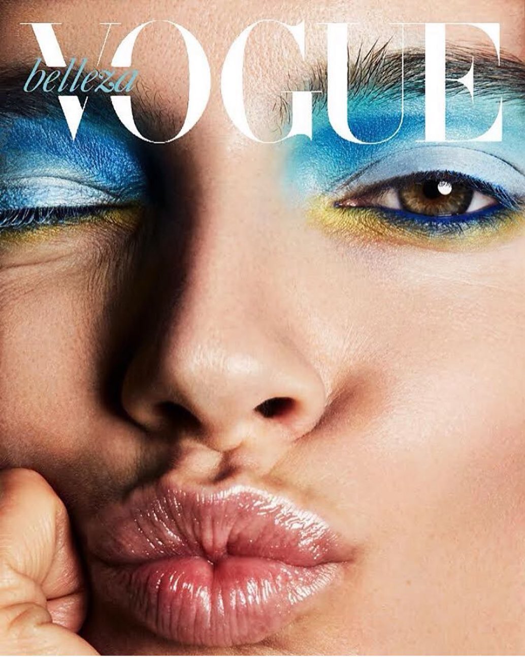 Close-up portrait of a woman's face on a magazine cover, featuring bright blue and yellow eye makeup and glossy lips.