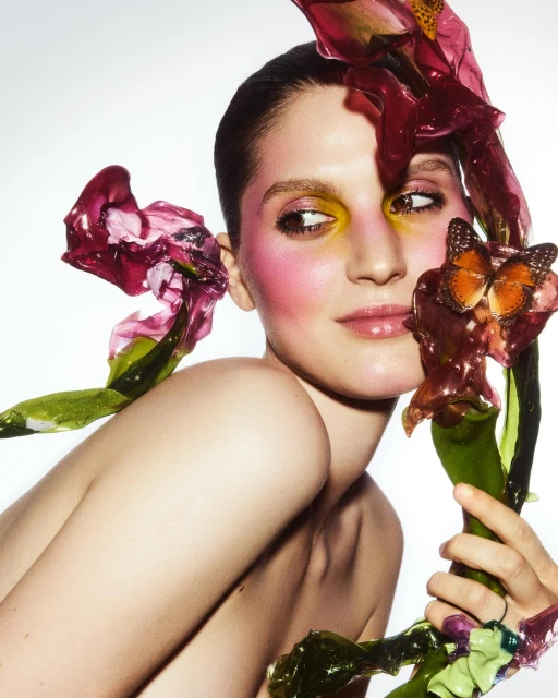 Close-up portrait of a woman with yellow and pink makeup, adorned with flowers and a butterfly, set against a bright white backdrop.