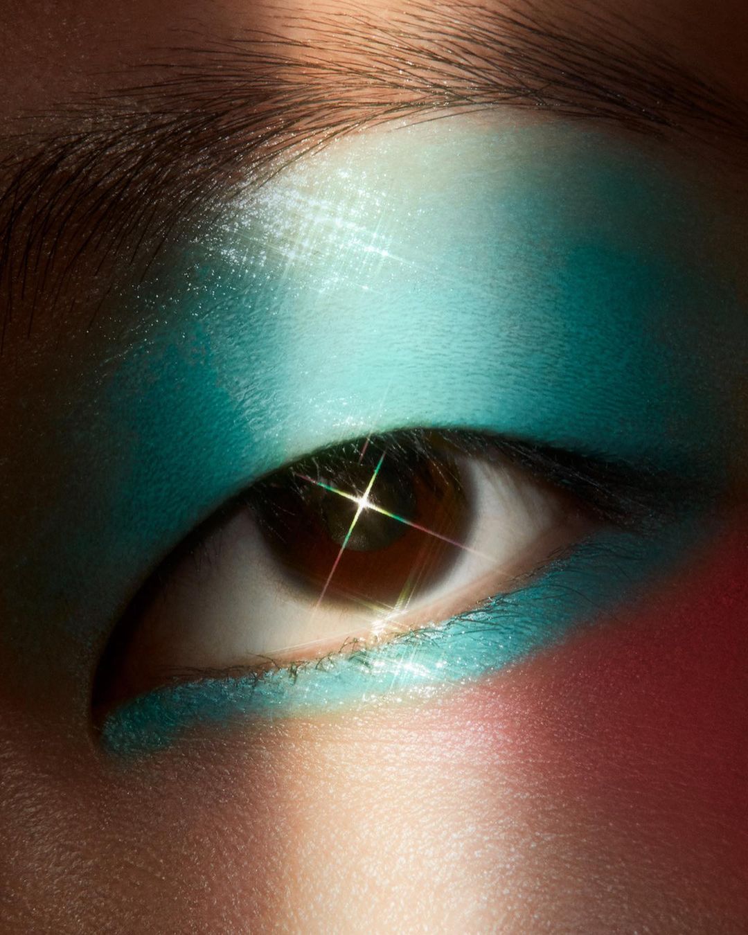 Macro shot of a brown eye with bright turquoise eyeshadow applied to the eyelid and lower lash line with a starburst lens flare.
