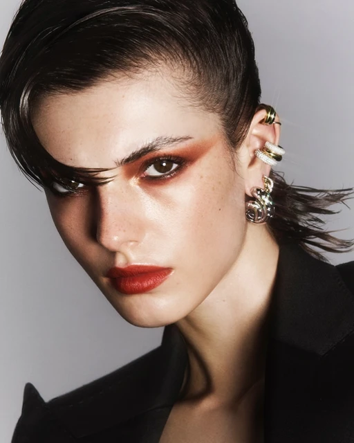 Close-up portrait of a woman with red lipstick and dramatic eye makeup wearing a black blazer against a neutral grey background.