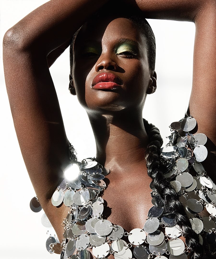Close-up studio shot of a Black female model in silver disc dress, green eye shadow, and glossy lips with white background and soft lighting.