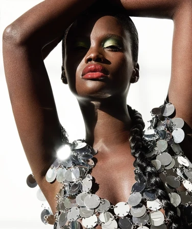 Close-up studio shot of a Black female model in silver disc dress, green eye shadow, and glossy lips with white background and soft lighting.