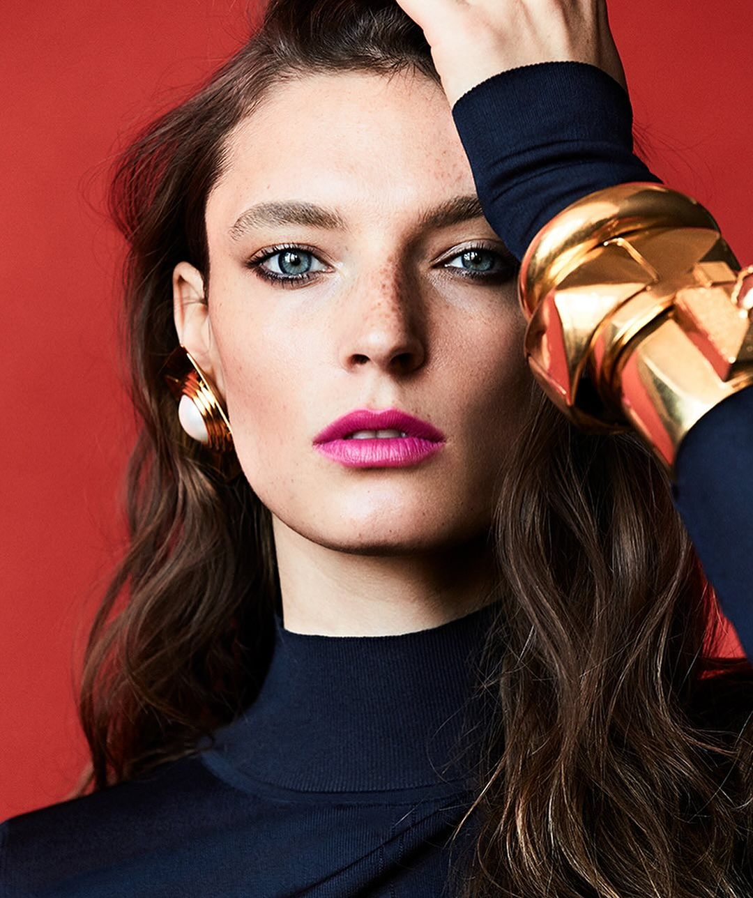 Close-up portrait of a woman wearing chunky gold bracelets, oversized earrings, and a dark top against a vibrant red background.
