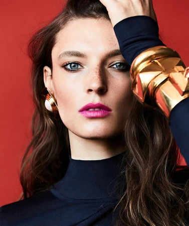Close-up portrait of a woman wearing chunky gold bracelets, oversized earrings, and a dark top against a vibrant red background.