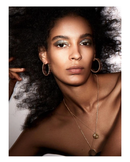 Close-up of a woman with dark curly hair, sparkling eye makeup, gold hoop earrings, and layered gold necklaces against a neutral background.