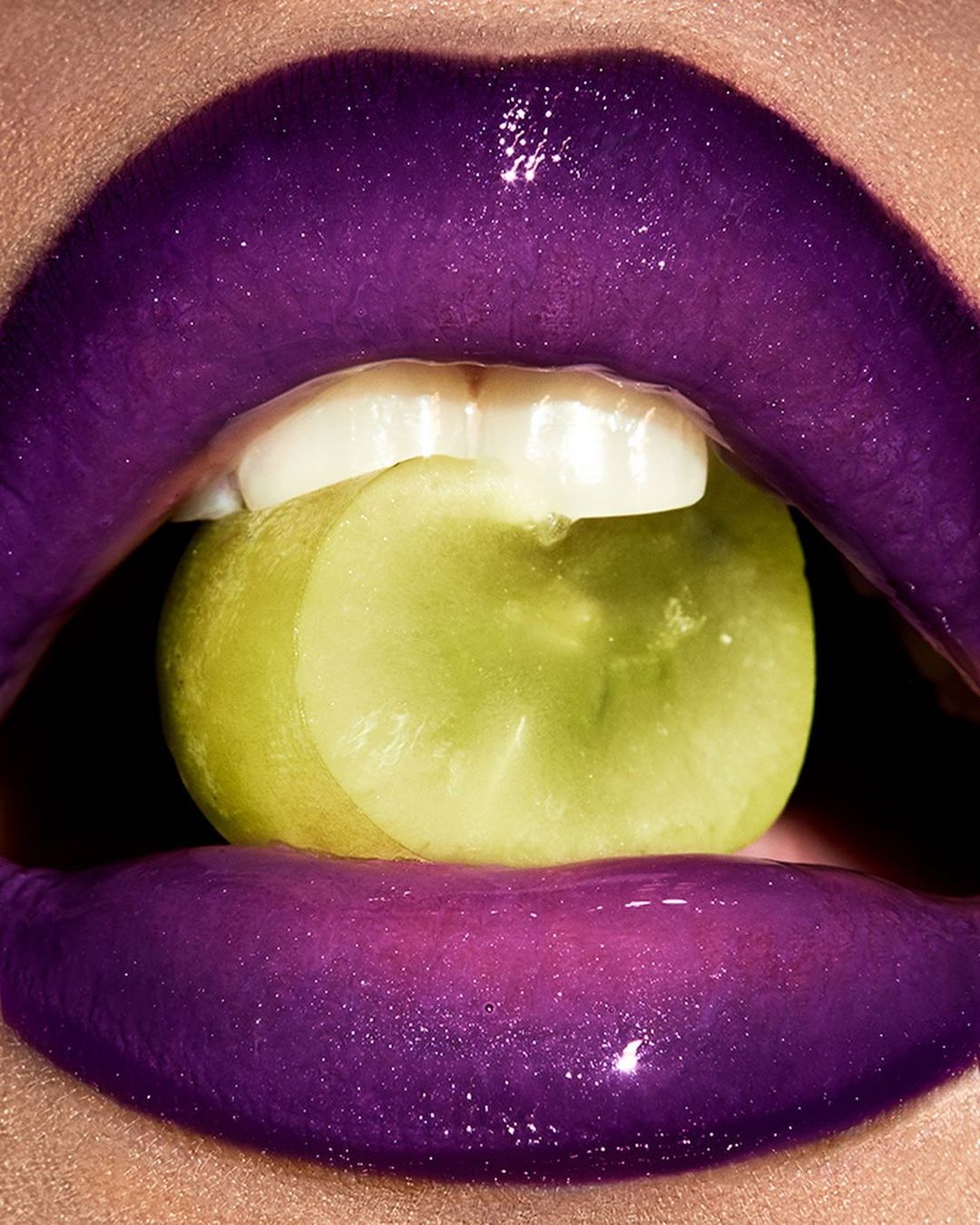 Close-up of lips with shiny purple lipstick, holding a green grape between teeth. Beauty shot for makeup and cosmetic product.