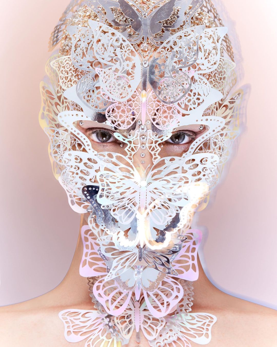 Close-up portrait of a person wearing a silver-and-white decorative butterfly mask and collar, with a light pink background.