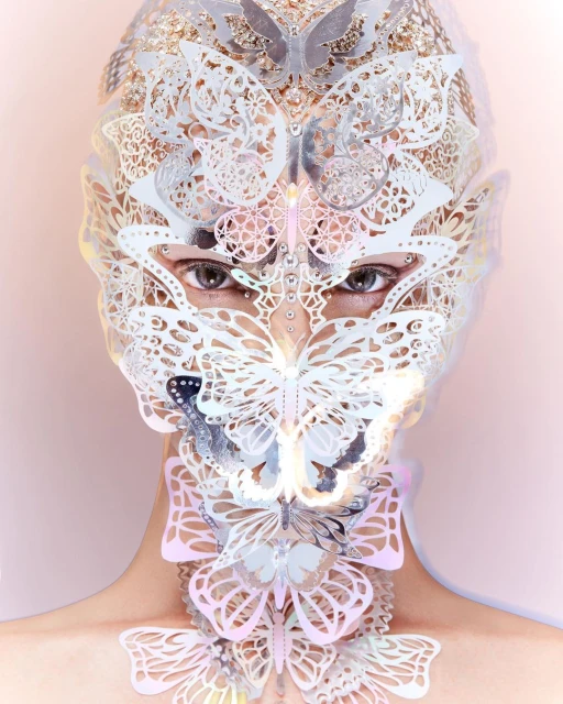 Close-up portrait of a person wearing a silver-and-white decorative butterfly mask and collar, with a light pink background.