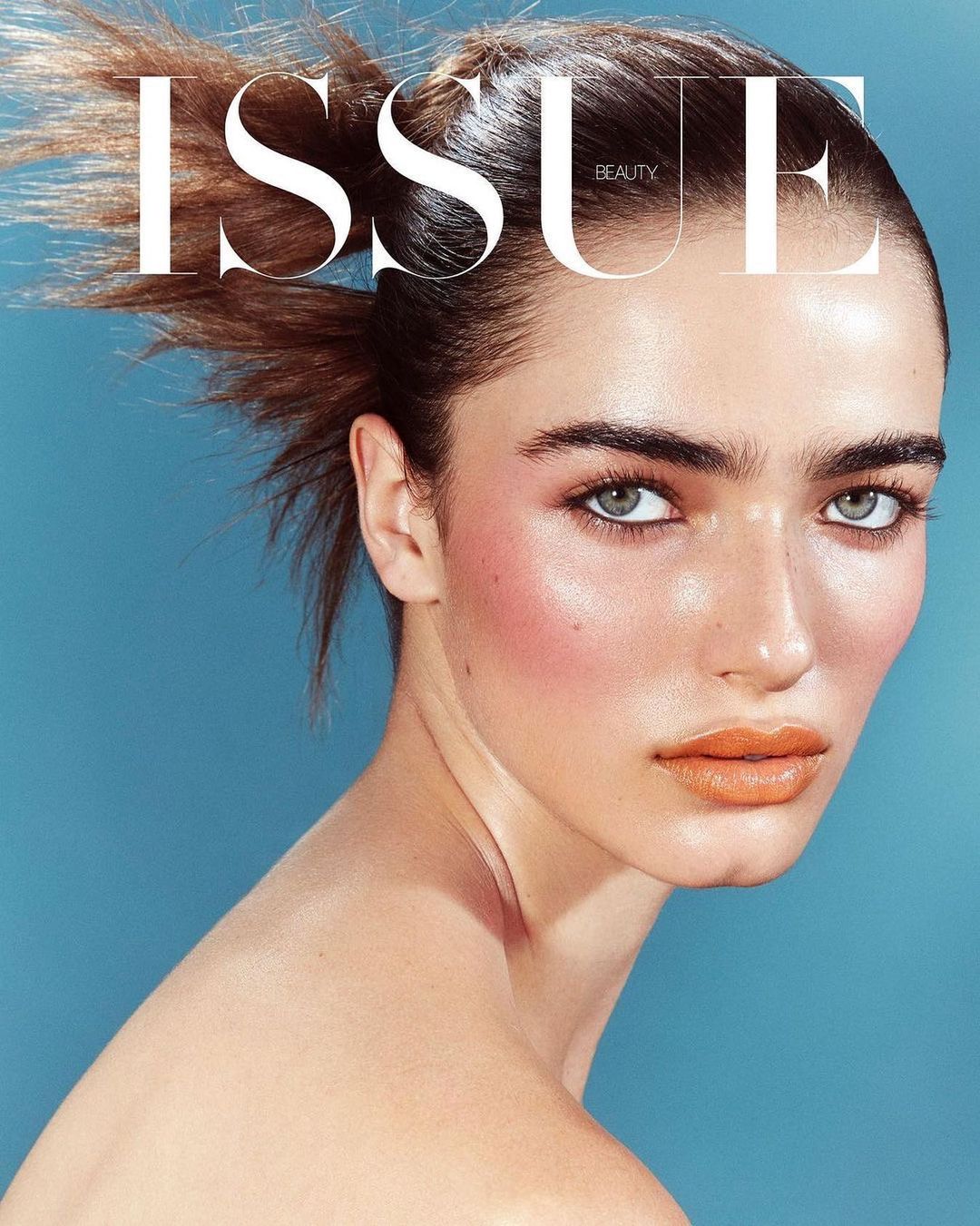 Close-up beauty shot of a woman with fair skin, soft makeup, and wind-blown hair on a blue background; beauty magazine cover.