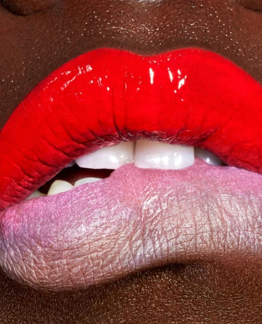 Close-up of red and pink glossy lips with teeth visible, dark skin tone, beauty shot emphasizing makeup and detail, with a focus on texture.