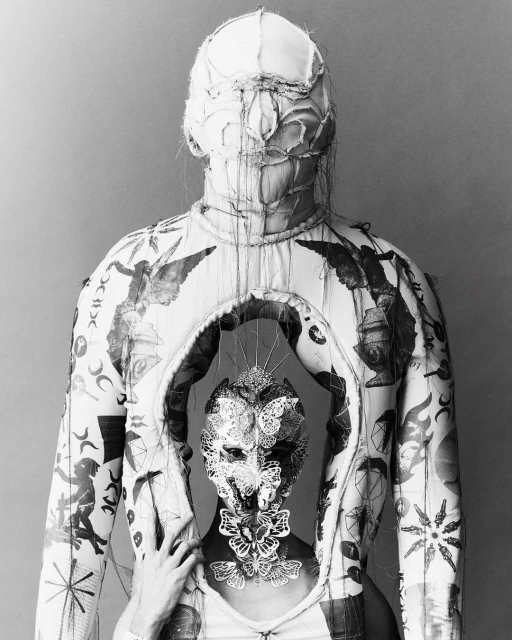 Monochrome image featuring person with a sculptural headpiece and garment adorned with applied art, captured in a studio setting.