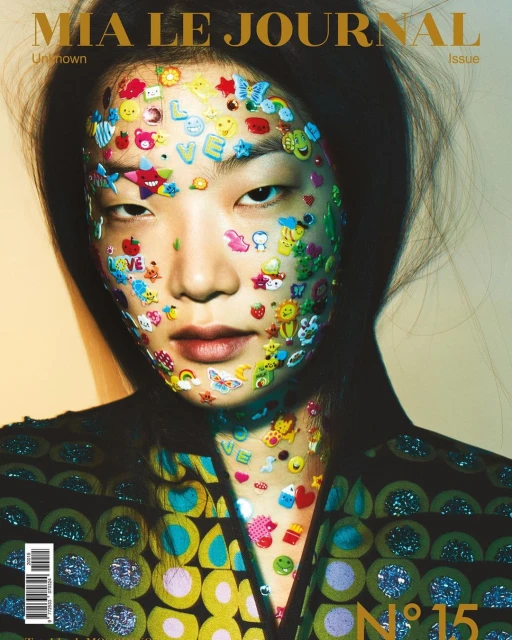 Magazine cover featuring an Asian woman with colorful stickers on her face and geometric-patterned jacket, creating a unique and artistic portrait.