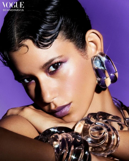 Beauty shot of a woman with short black hair wearing large silver statement earrings, bracelet, and purple eye makeup and lipstick.