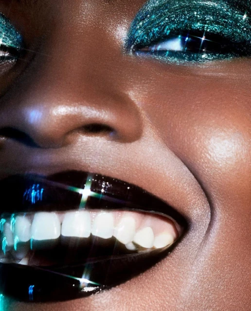 Close-up of a woman with emerald glitter eye makeup and dark glossy lips smiling to reveal white teeth; beauty and fashion.