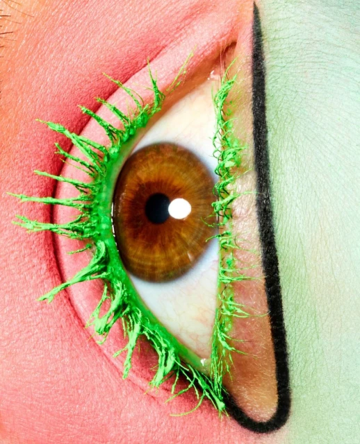 Close-up of a human eye featuring long, vibrant green eyelashes and graphic black eyeliner, set against a split peachy-pink and light green background.