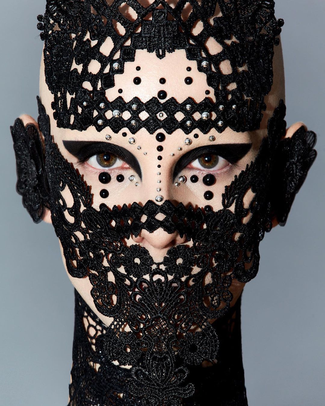 Striking close-up portrait of a bald adult female with black lace headpiece, dramatic eye makeup, and decorative beads against a grey backdrop.