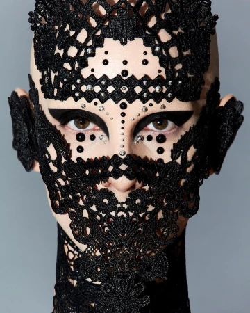 Striking close-up portrait of a bald adult female with black lace headpiece, dramatic eye makeup, and decorative beads against a grey backdrop.