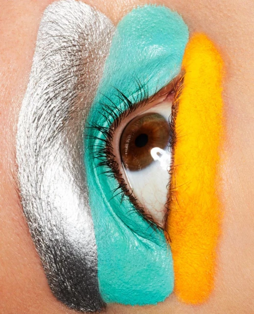 Close-up of a human eye with bright, colorful makeup of silver, aqua, and yellow stripes applied around the eye for a vibrant artistic look.