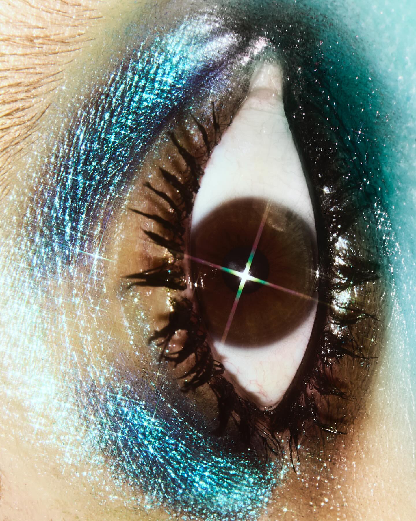 Close-up of a single eye with dramatic teal and blue eye shadow. The eye has dark lashes and a bright starburst reflection.