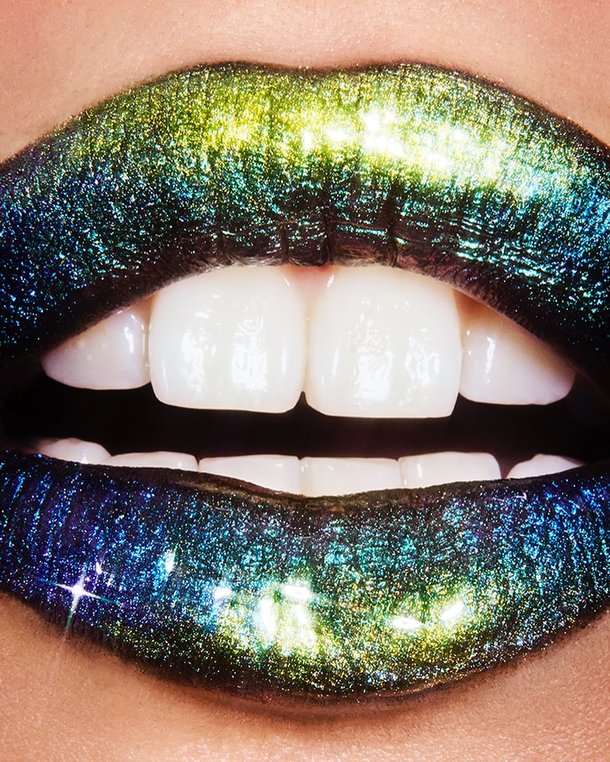Extreme close up of lips wearing shimmering metallic green and blue glitter lipstick showing white teeth