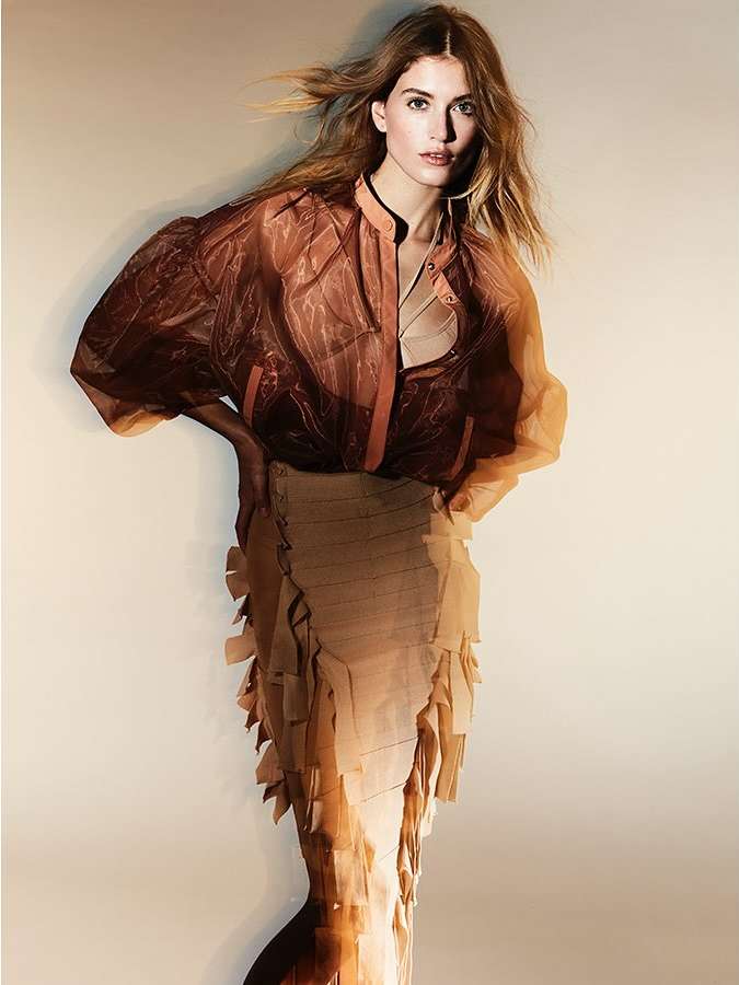 Portrait of a young adult woman posing in a semi-sheer brown blouse and textured tan skirt against a neutral background in a studio.