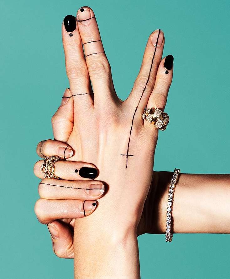 Woman's hands with black nail polish and graphic black lines and shapes adorn fingers, featuring jewelry rings and bracelet on blue.
