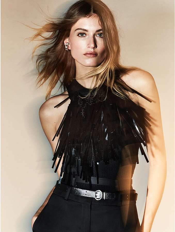 A woman in black high-waisted pants, a black top, and a fringed halter neck with a belt, poses in front of a neutral background.