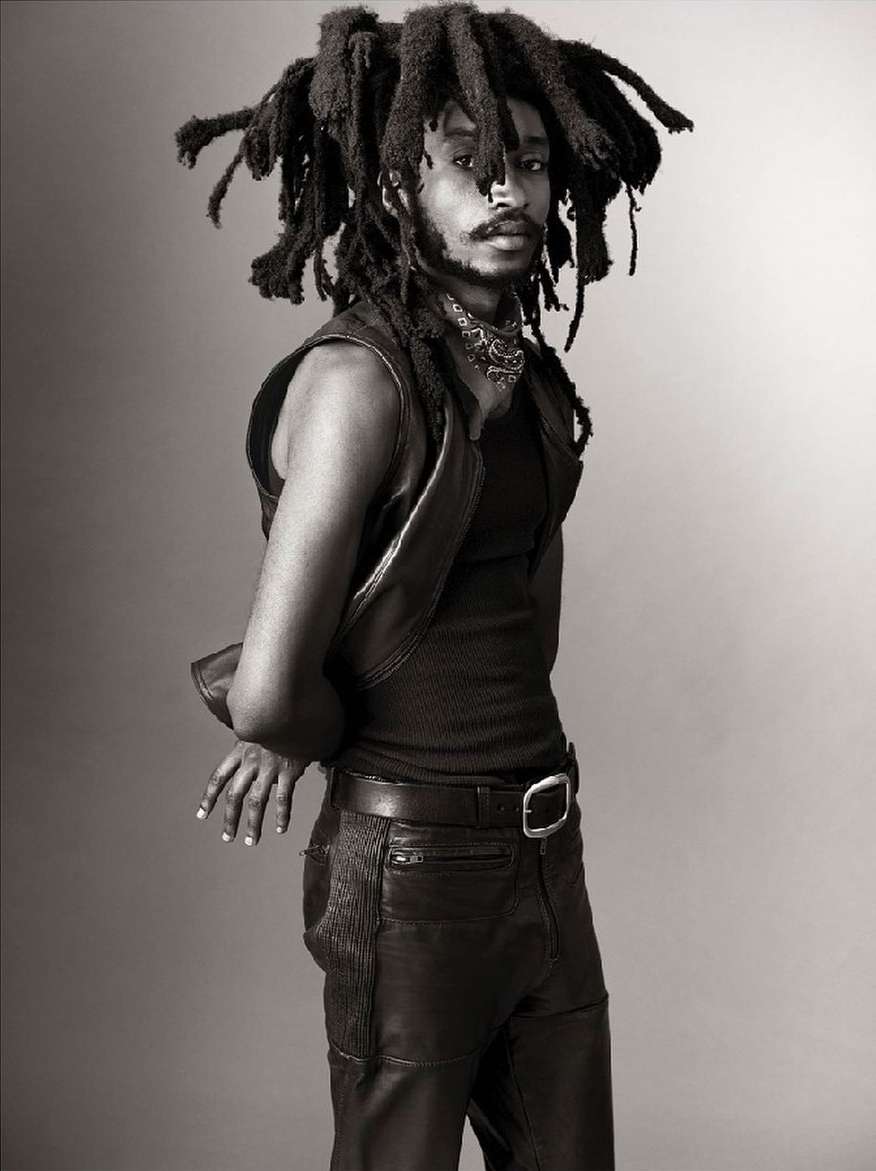 Portrait of adult male with dreadlocks wearing a vest and leather pants against a grey backdrop in a studio setting.