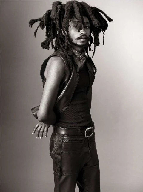 Portrait of adult male with dreadlocks wearing a vest and leather pants against a grey backdrop in a studio setting.
