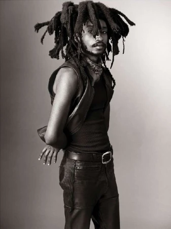 Portrait of adult male with dreadlocks wearing a vest and leather pants against a grey backdrop in a studio setting.