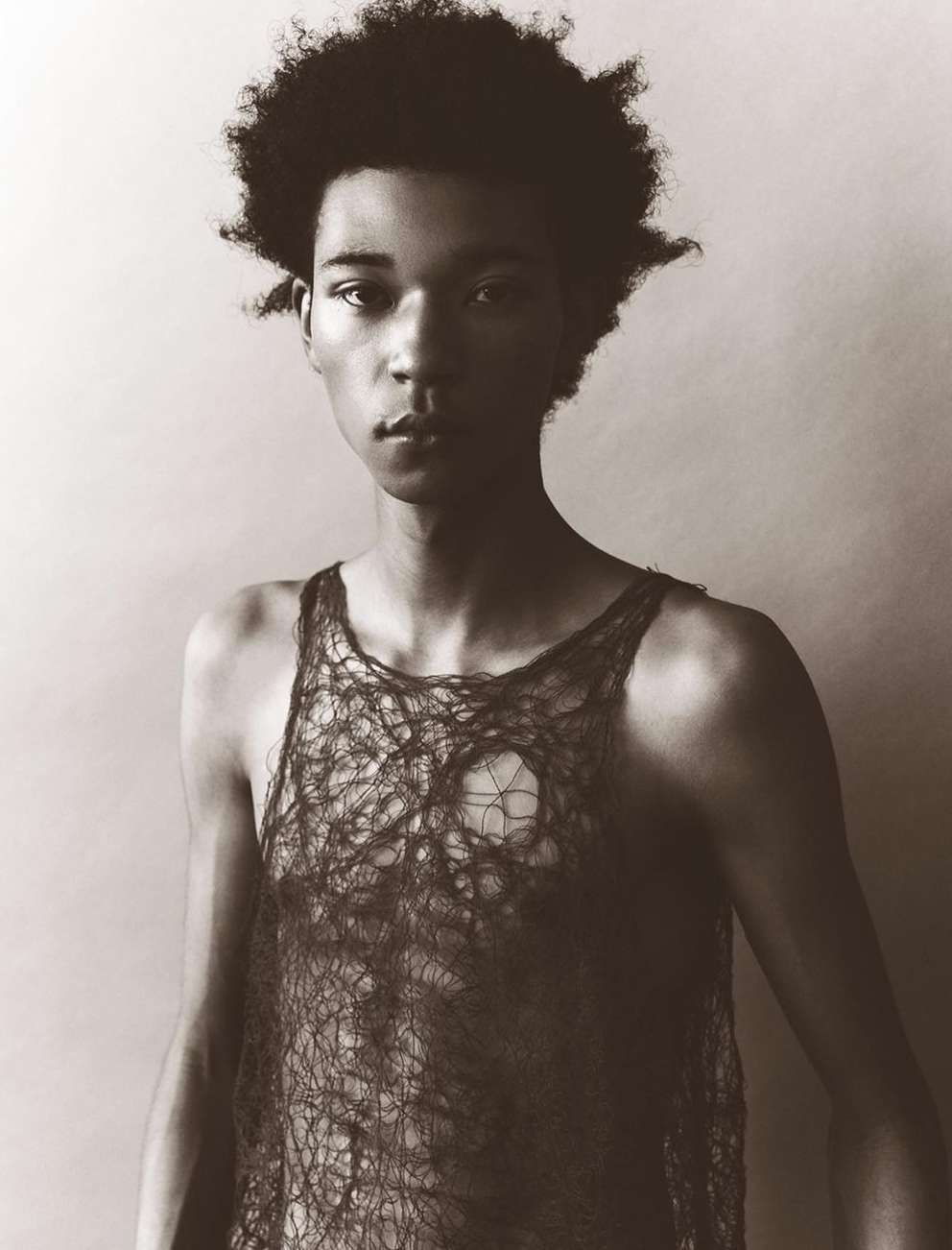 Portrait of a young Black man with curly hair in a see-through top, posing in a photo studio with a plain backdrop and subdued lighting.