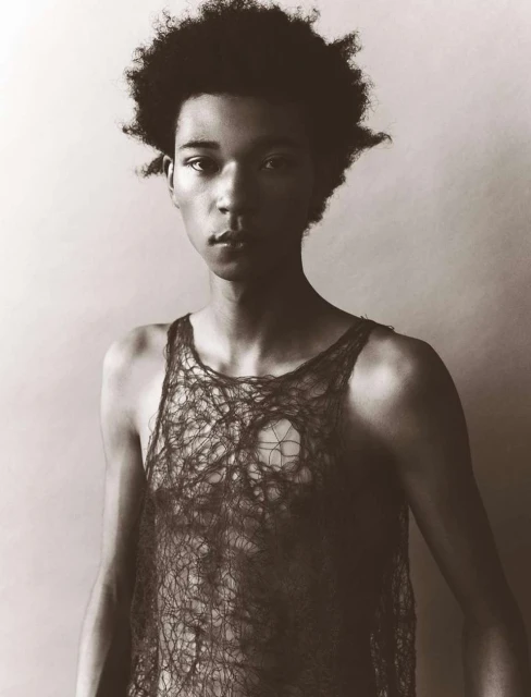 Portrait of a young Black man with curly hair in a see-through top, posing in a photo studio with a plain backdrop and subdued lighting.