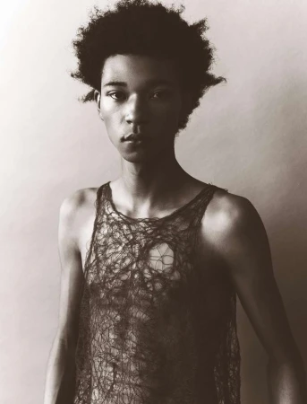 Portrait of a young Black man with curly hair in a see-through top, posing in a photo studio with a plain backdrop and subdued lighting.