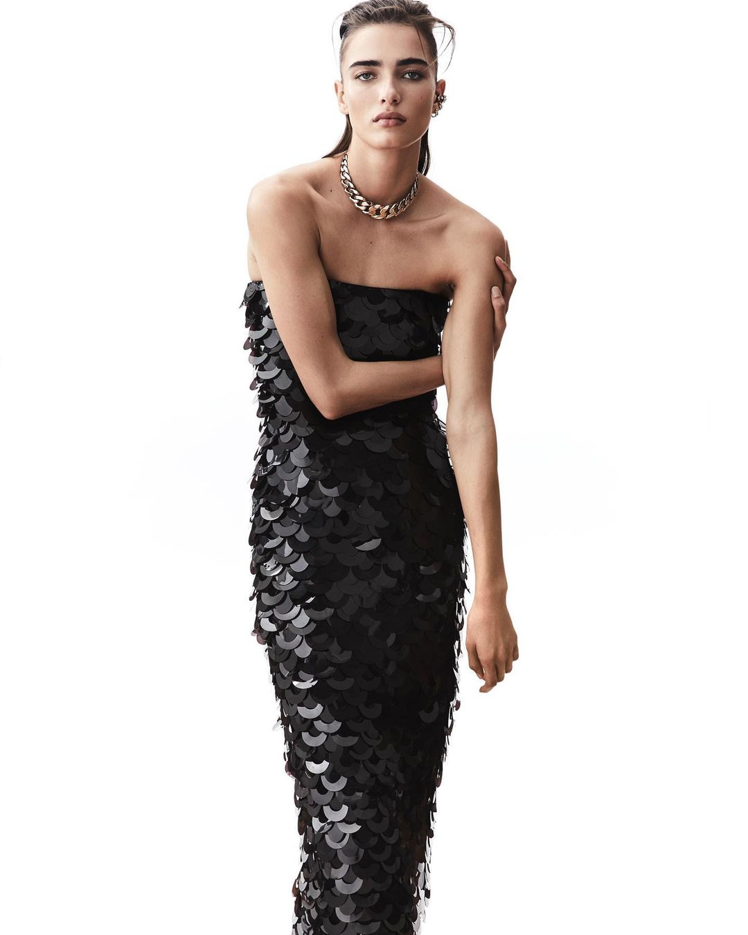 Elegant woman in black strapless sequin gown with chain necklace posing against a white background in a studio fashion shot.