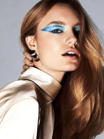 Close-up portrait of a woman with vibrant blue winged eyeshadow and long brown hair wearing a satin high-neck top by Mikael Schulz.
