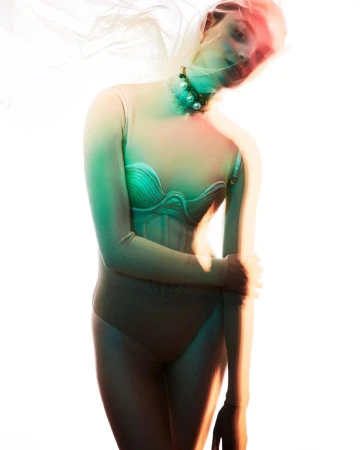 A woman wearing a bra top, bodysuit and pearl necklace is posing in a studio with soft lighting and a plastic wrap over her head.