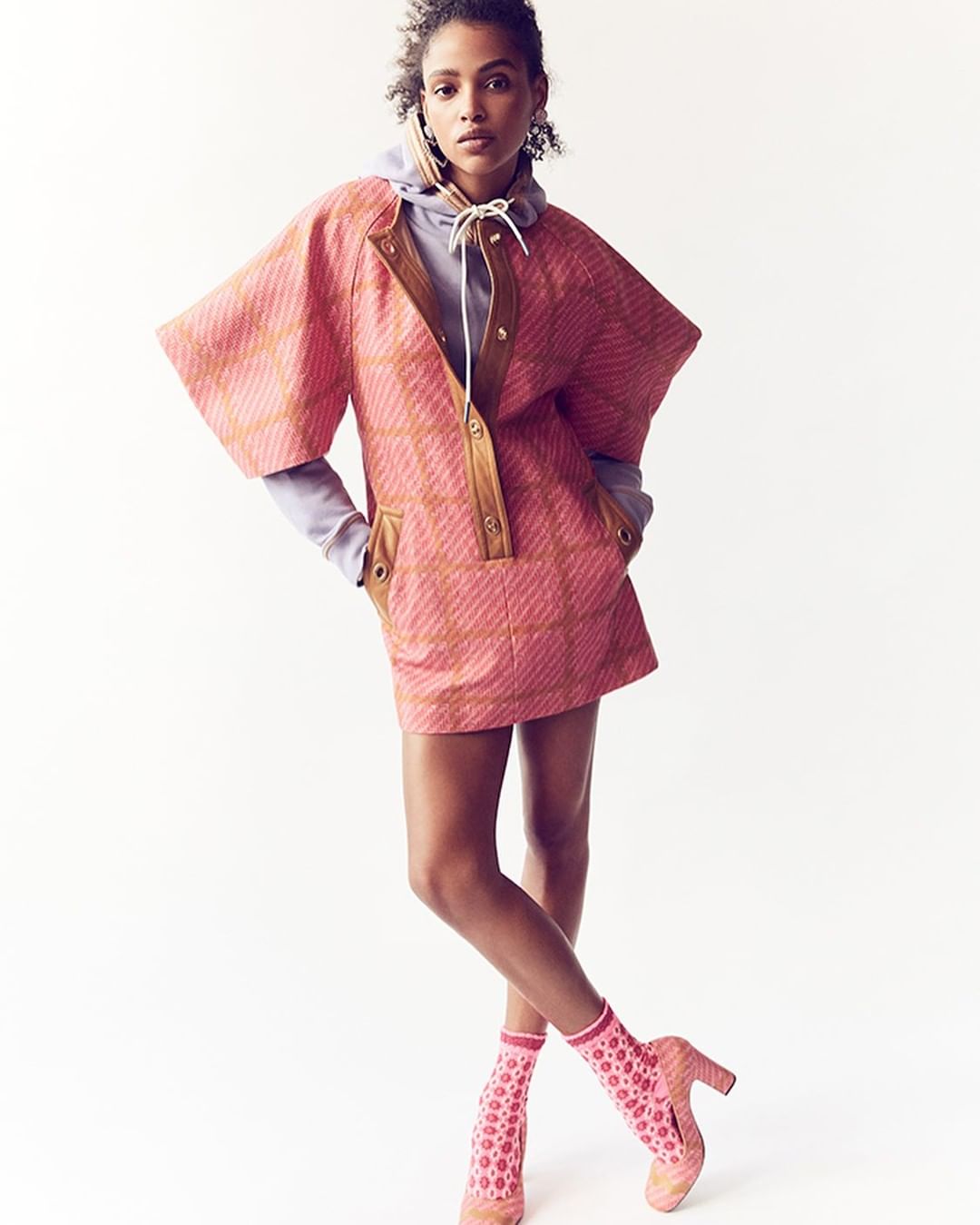 Editorial fashion shot of a Black woman posing in a pink patterned dress, lilac hoodie, and patterned shoes against a white background.