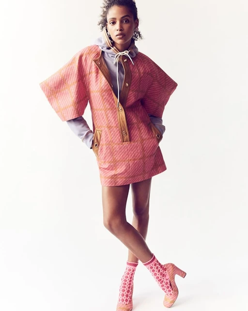 Editorial fashion shot of a Black woman posing in a pink patterned dress, lilac hoodie, and patterned shoes against a white background.