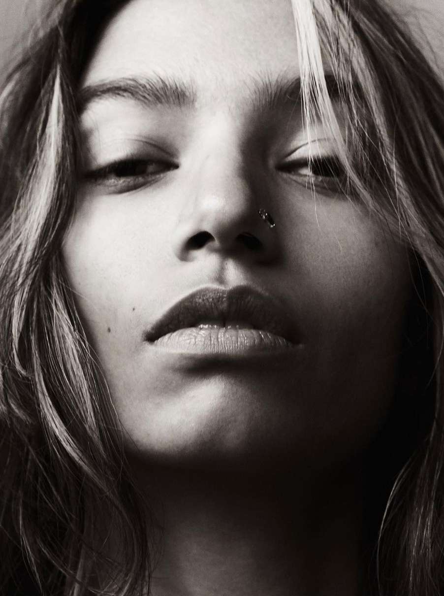 Close-up portrait of a young adult female with long hair, nose piercing, and neutral facial expression, captured in black and white with soft lighting.