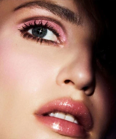 Close-up portrait of a woman featuring shimmering pink eyeshadow, glossy lips, and dark eyelashes against a dark background.