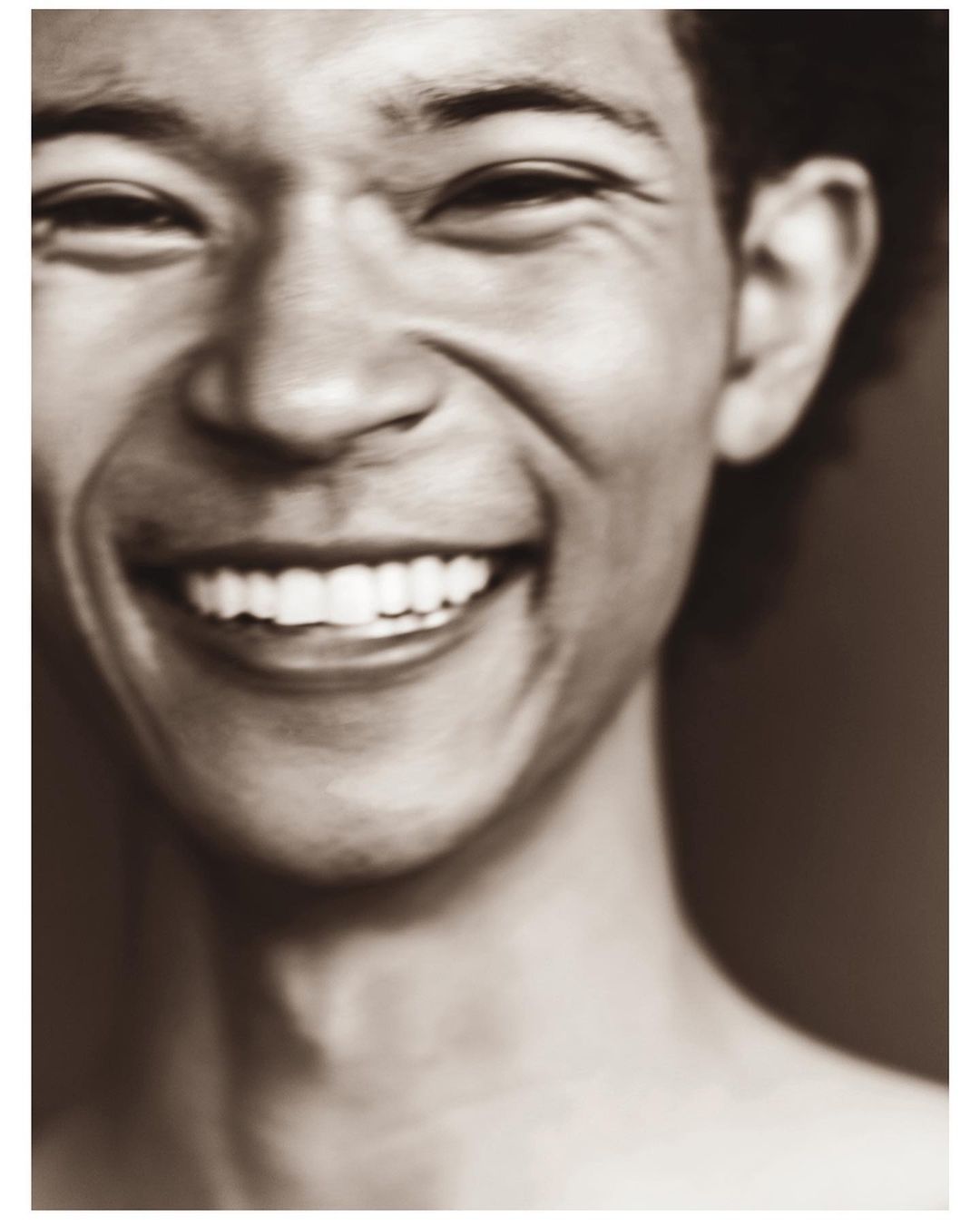 Close-up portrait of a smiling Asian man with fair skin showing his teeth, against a blurred sepia-toned background.