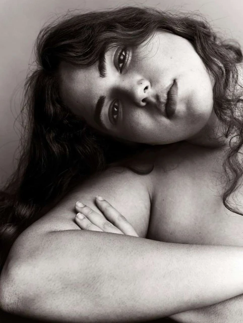 Black and white portrait of an adult woman with long, dark curly hair gazing forward with her arms crossed in front of her body.