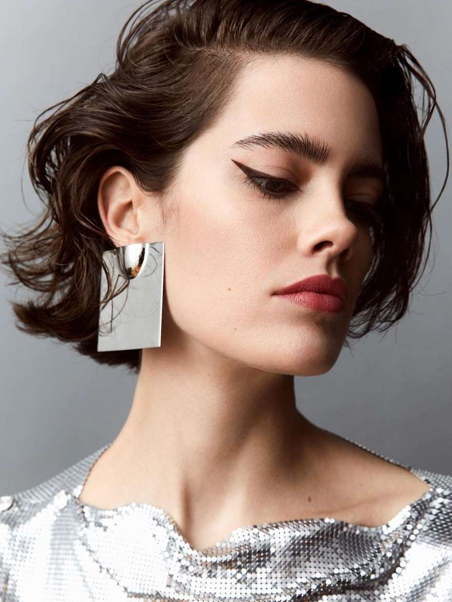 Close-up studio shot of a woman with short wavy dark hair, wearing a silver sequin top and large rectangular reflective silver earrings.