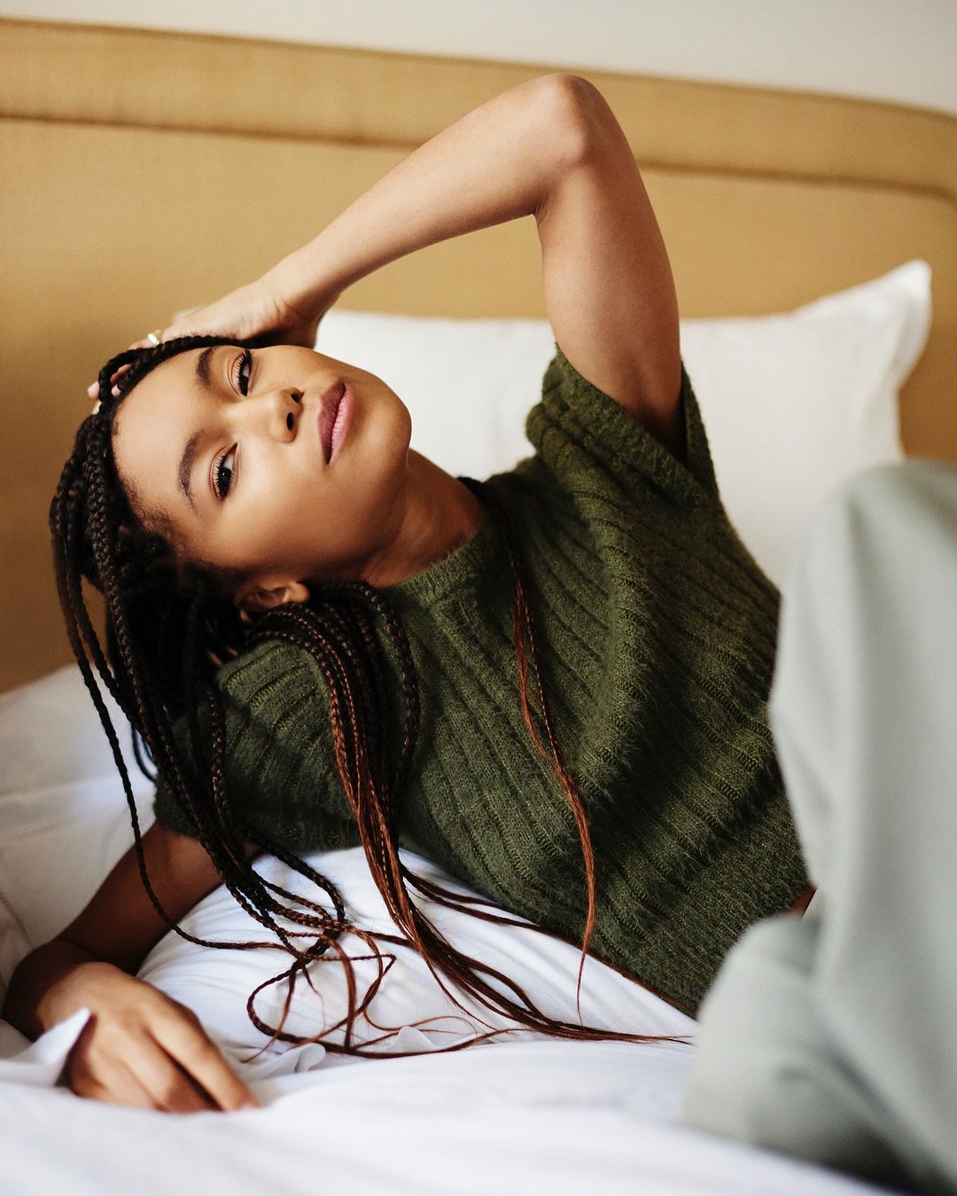 Portrait of a Black woman with braided hair lying on a bed in a green sweater, looking at the camera with a relaxed expression.